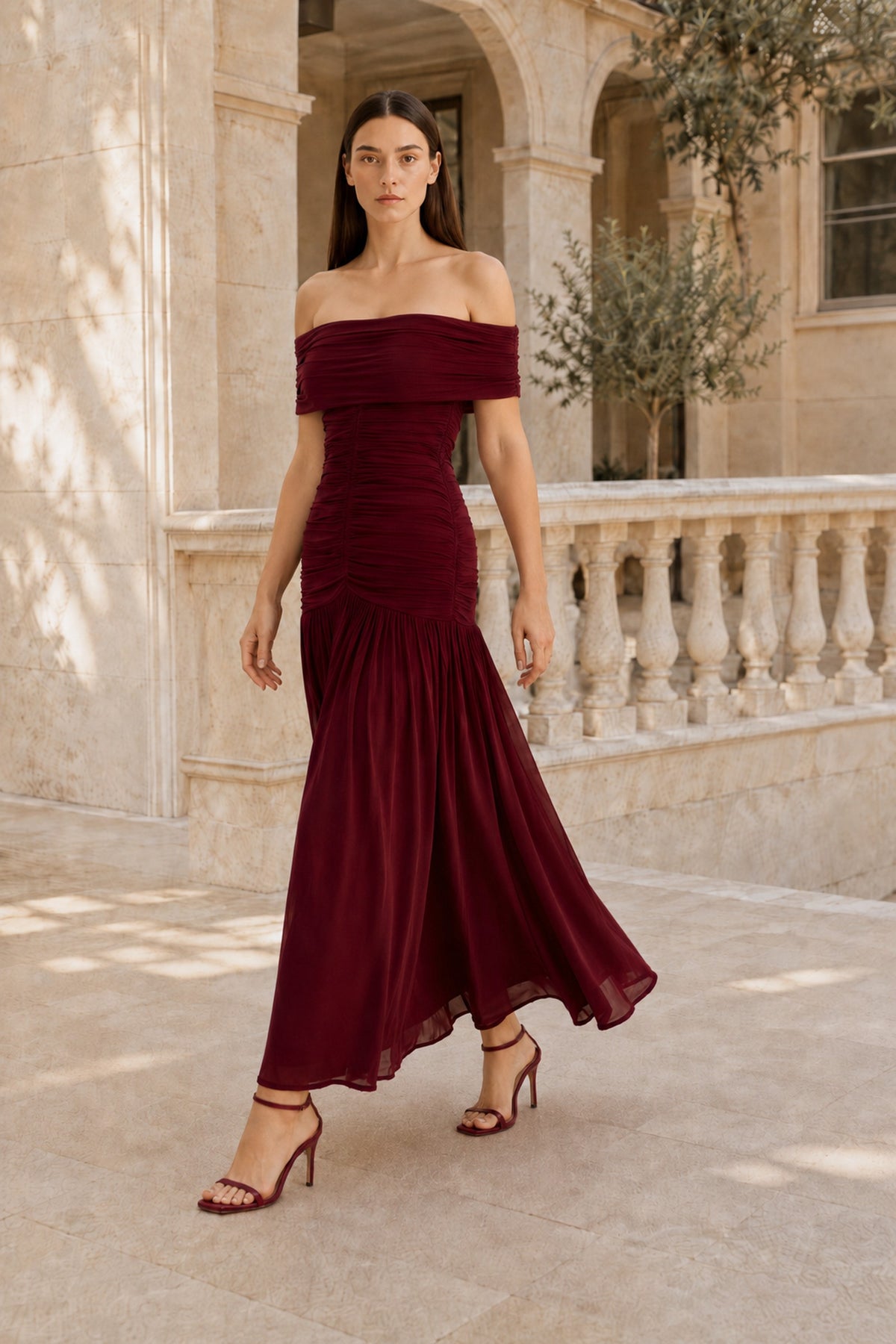 Camille burgundy off-shoulder dress shown in motion, with a ruched bodice and flowing skirt in a stone courtyard