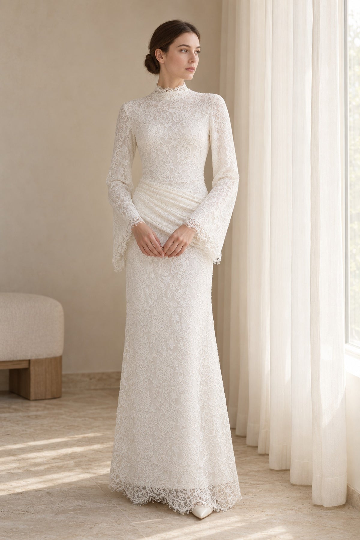 Lilia white lace dress on a model beside a window, showing the high neckline, long sleeves and ruched hip panel