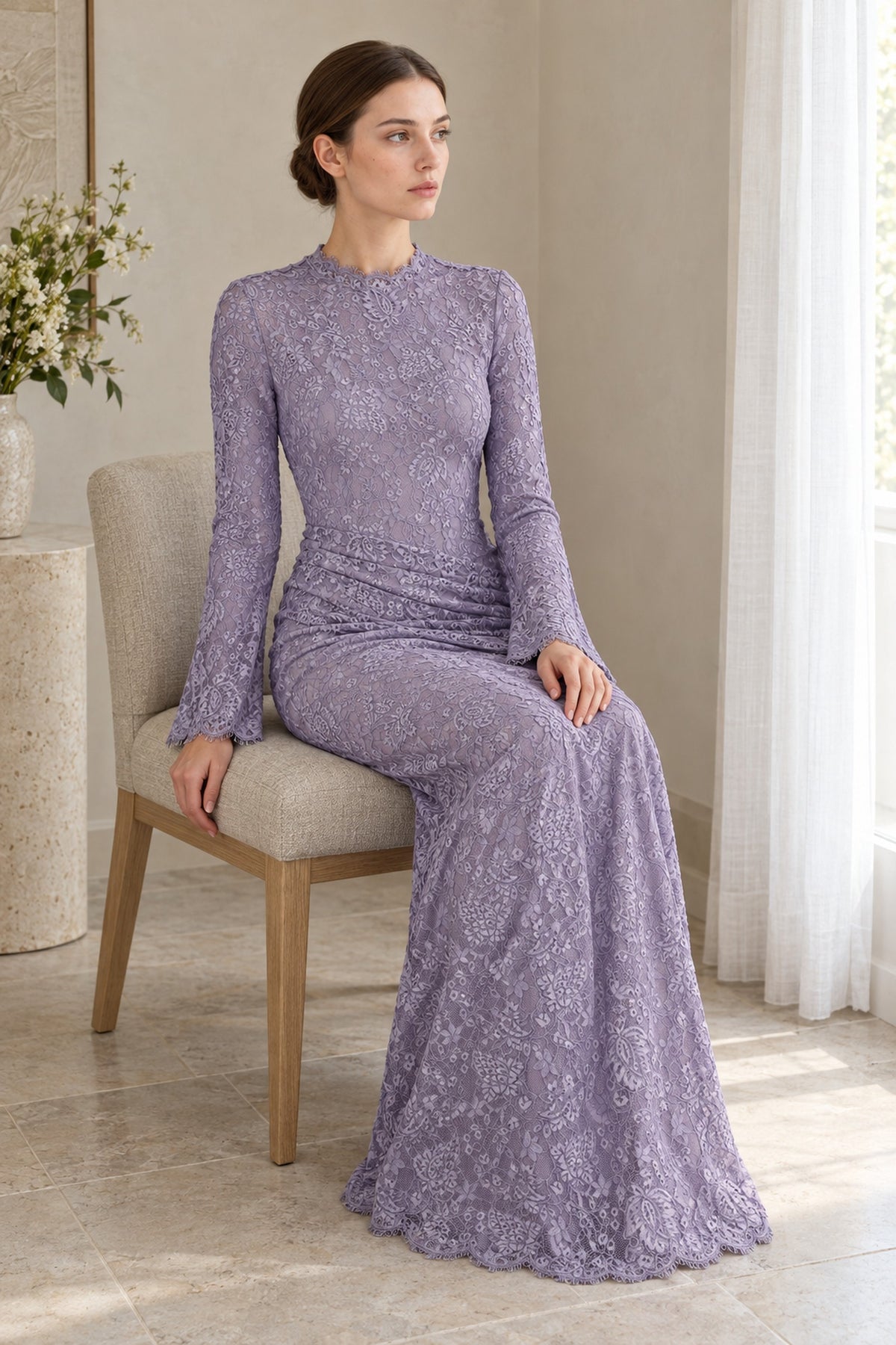 Seated model wearing the Lilia lilac lace maxi dress by a window, showing the waist drape and scalloped hem.