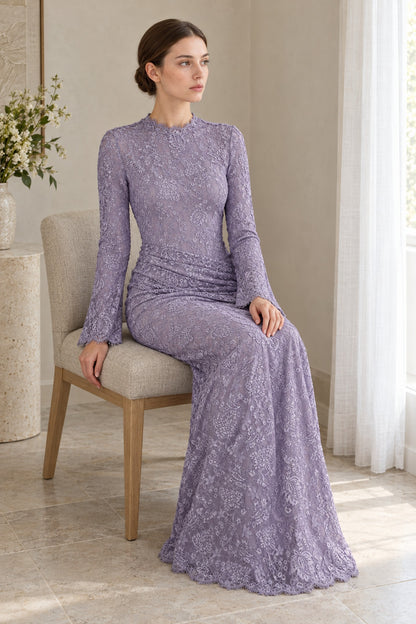 Seated model wearing the Lilia lilac lace maxi dress by a window, showing the waist drape and scalloped hem.